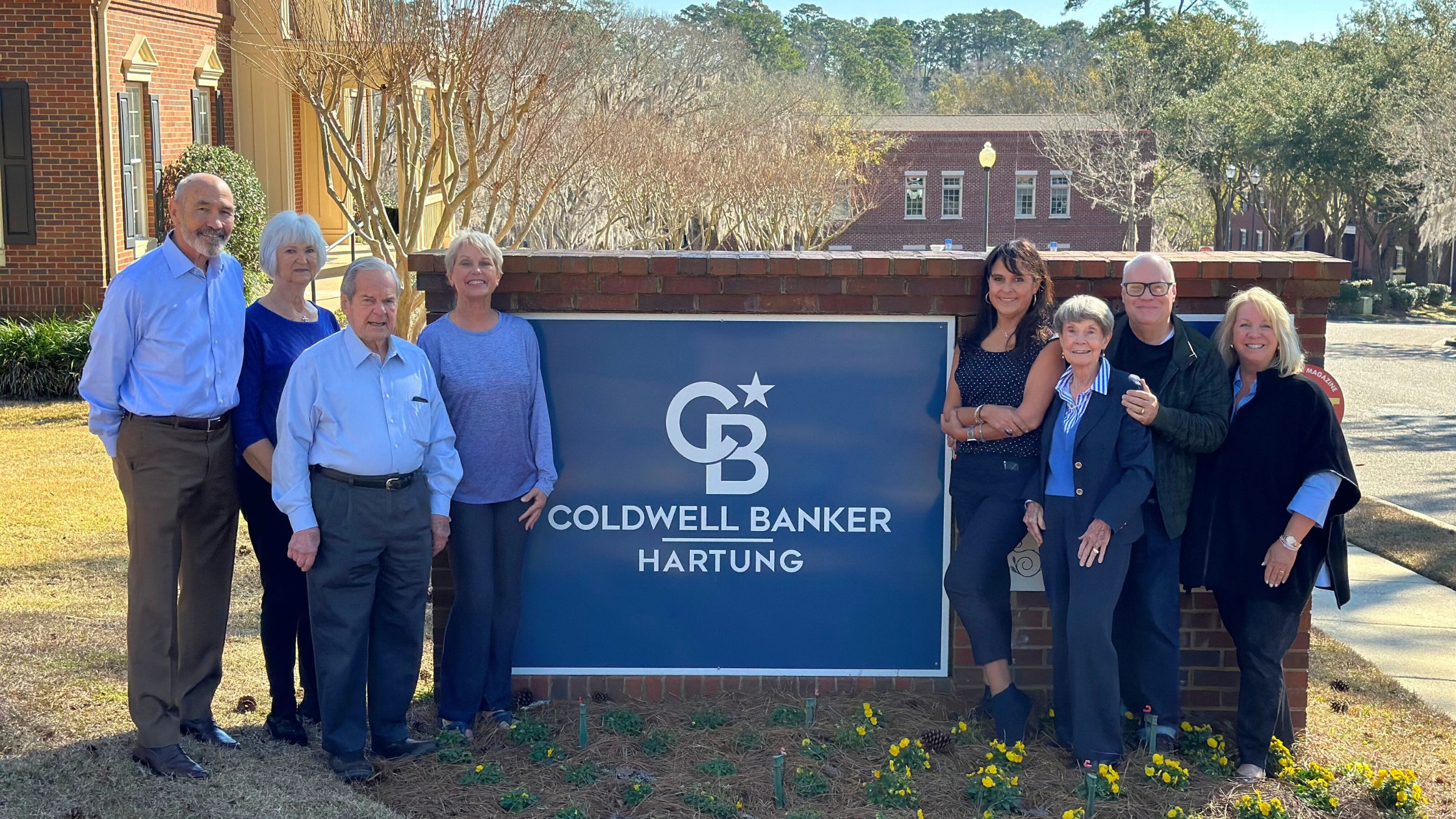A photo of 8 Realtors posing and smiling in front of the Coldwell Banker Hartung sign. They are all wearing blue shirts and/or jackets in various tones. There are 2 men and 2 women on the left of the sign, and 1 man and 3 women on the right. This photo was taken during the day with bright lighting.