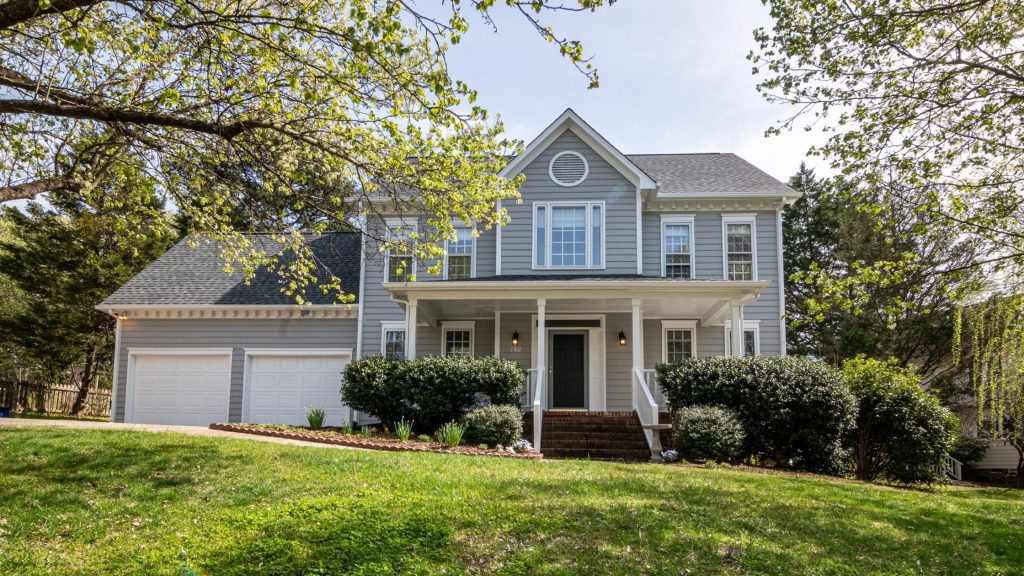 Stock photo of the exterior of a two-story home. The home is blue-grey with two garage doors on the left side and the main house on the right. It is sitting on green grass with perfectly manicured bushes in front og the main house. There are tree branches with green leaves peeking from the top corners of the photo.